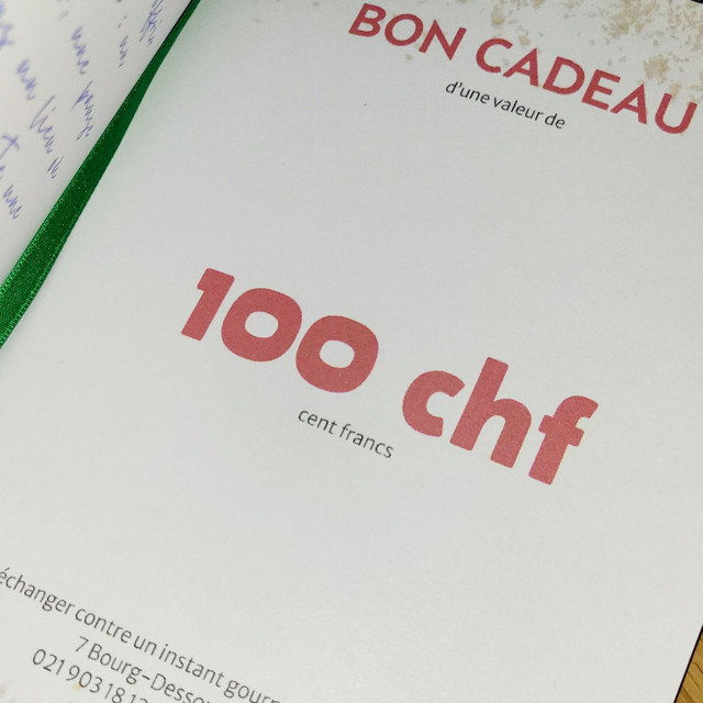 Certificat cadeau blanc à texte rouge, portant la mention « 100 CHF », inscription française « Bon Cadeau » en haut, reliure verte à gauche.