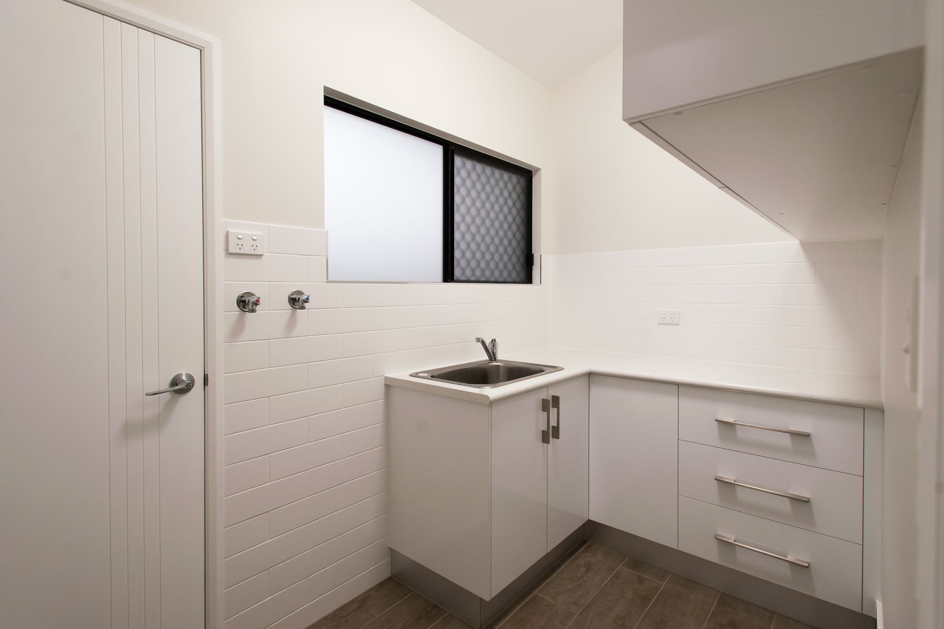 A Modern Laundry Room With White Cabinetry, A Stainless Steel Sink, Tiled Walls, And A Window — B & C Cabinets in Mareeba, QLD