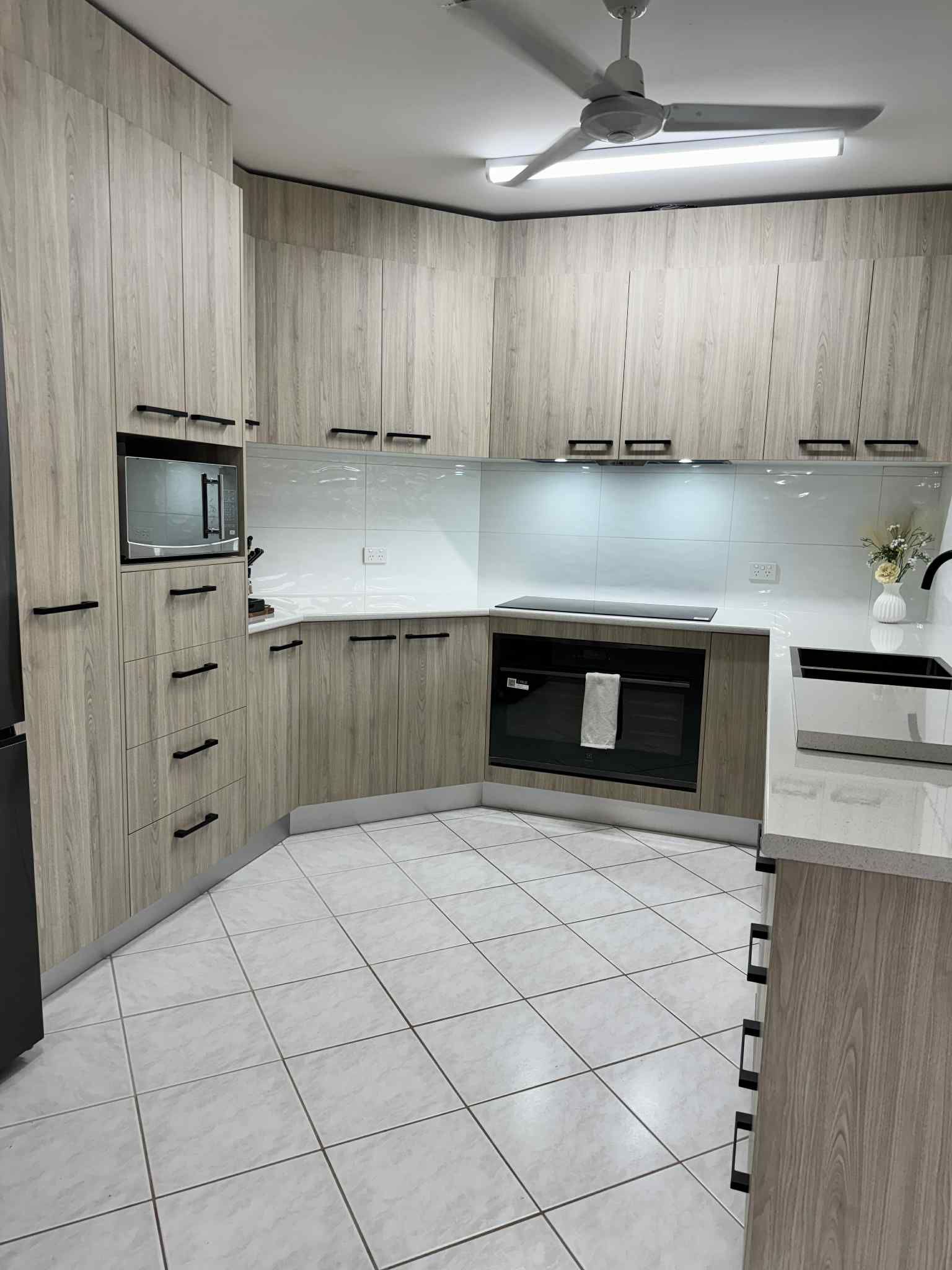A Modern Kitchen With Light Wood-grain Cabinets, White Countertops, Black Hardware, And White-and-grey Tiled Floors — B & C Cabinets in Mareeba, QLD