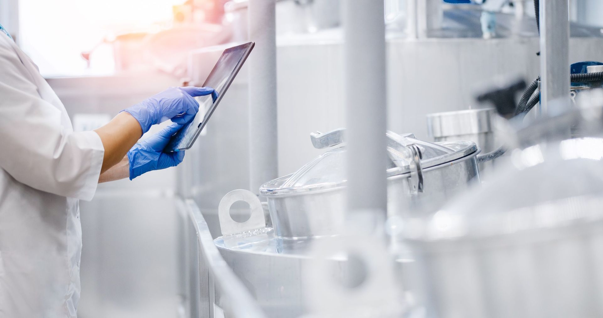 A person in a white lab coat and blue gloves uses a tablet while standing in a clean, industrial laboratory setting.