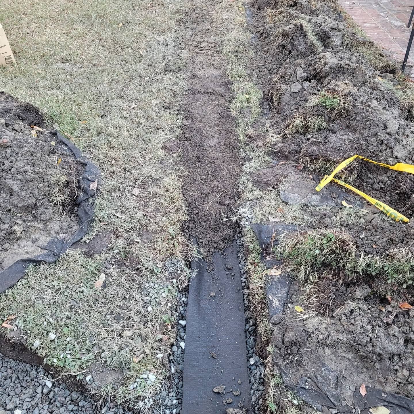 Narrow muddy trench in a grassy yard, bordered by soil piles and black plastic edging.