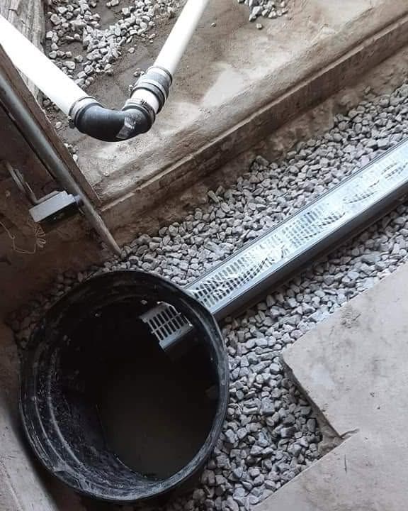Black bucket in gravel beside a perforated drain pipe and exposed plumbing under construction