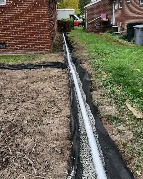 A white drainage pipe runs through a narrow trench beside a brick house and grassy yard.