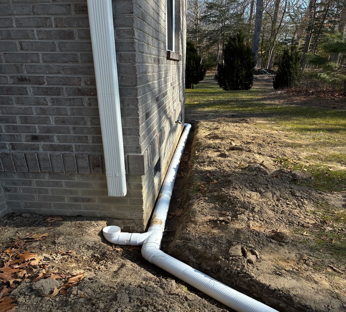 Brick house corner with white drainage pipe running along the ground into a yard