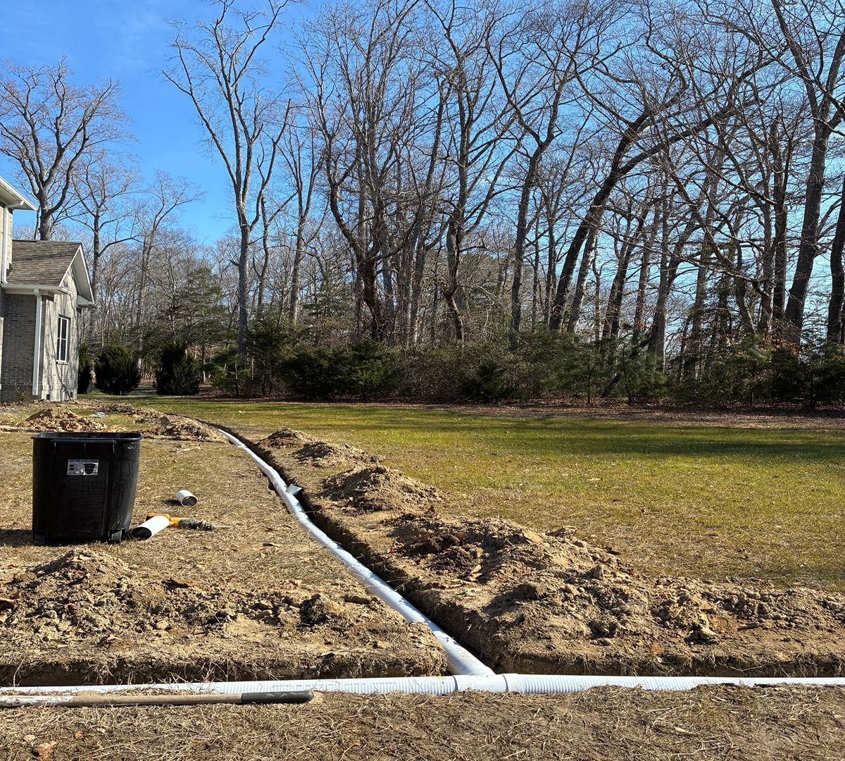 Backyard with a black trash bin, white pipes, and a dug trench beside a house under bare trees