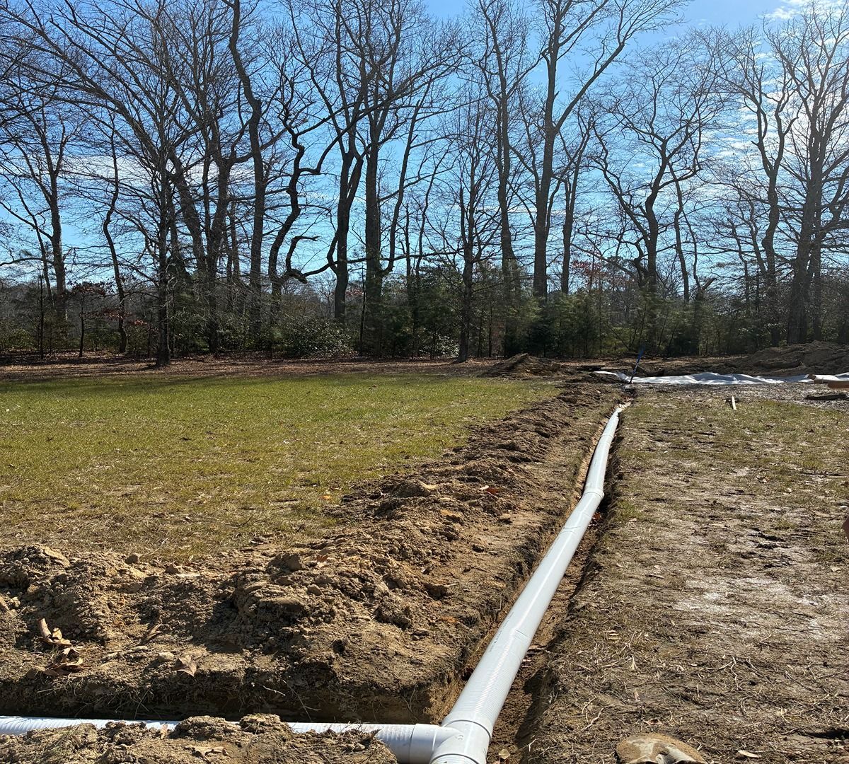 A white pipe runs through a muddy field beside a grassy area and leafless trees under a blue sky.