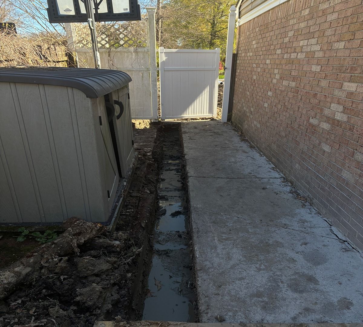 Narrow side yard between a brick wall and fence, with a gray storage shed and concrete path.
