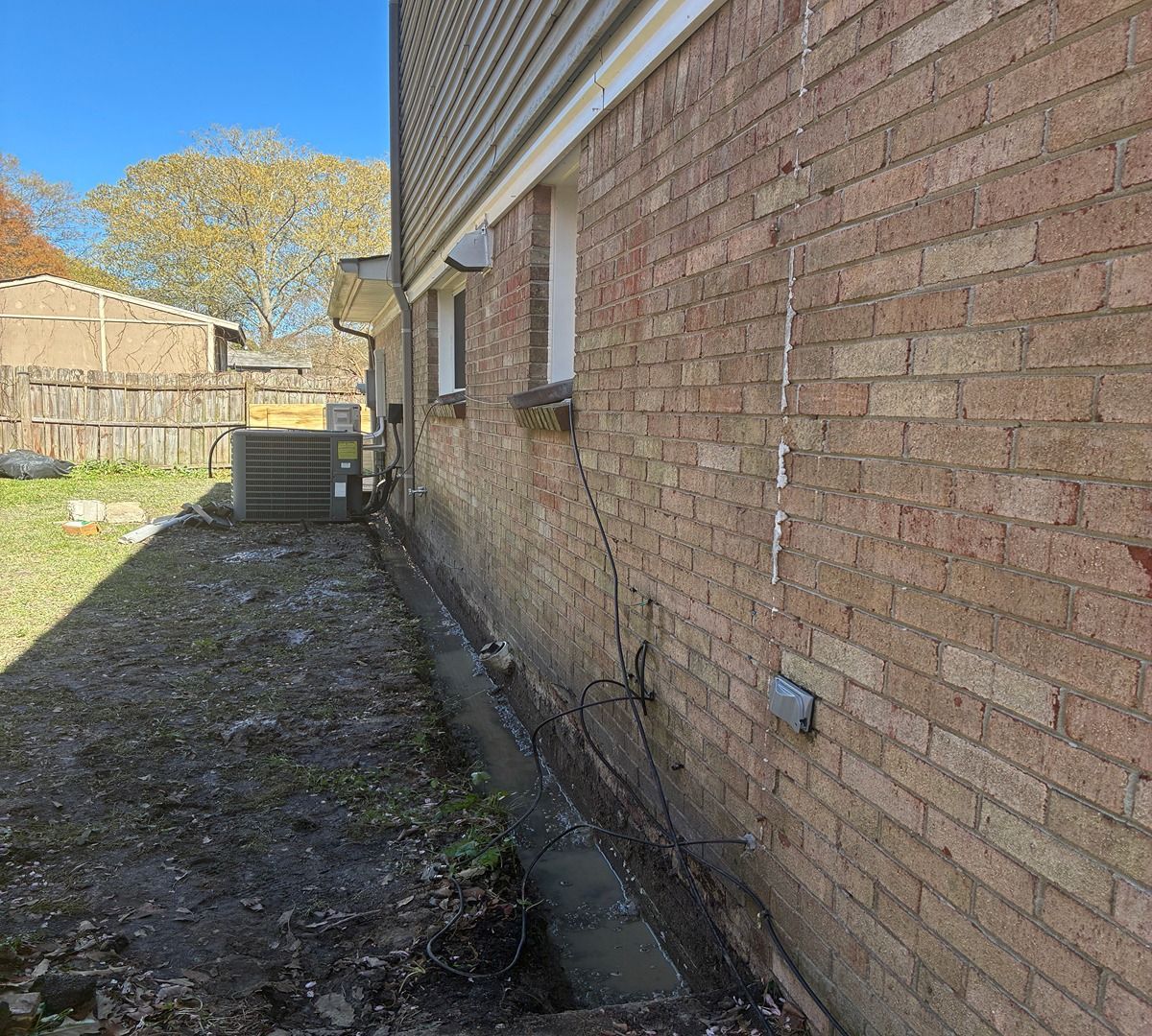 Narrow side yard beside a brick house, with a fence, dirt ground, and HVAC unit in the distance.