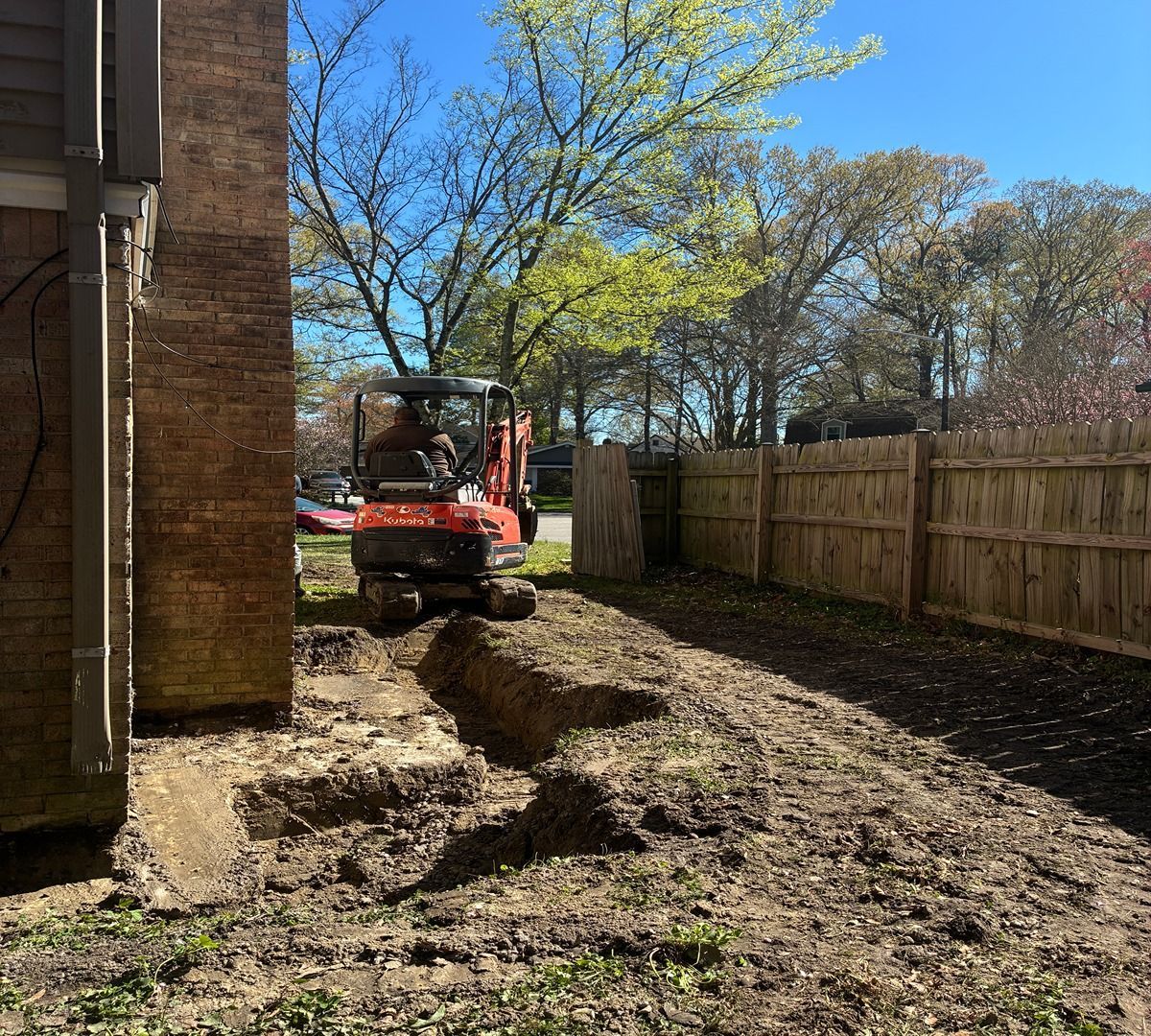 Mini excavator digging a trench beside a house in a fenced backyard on a sunny day