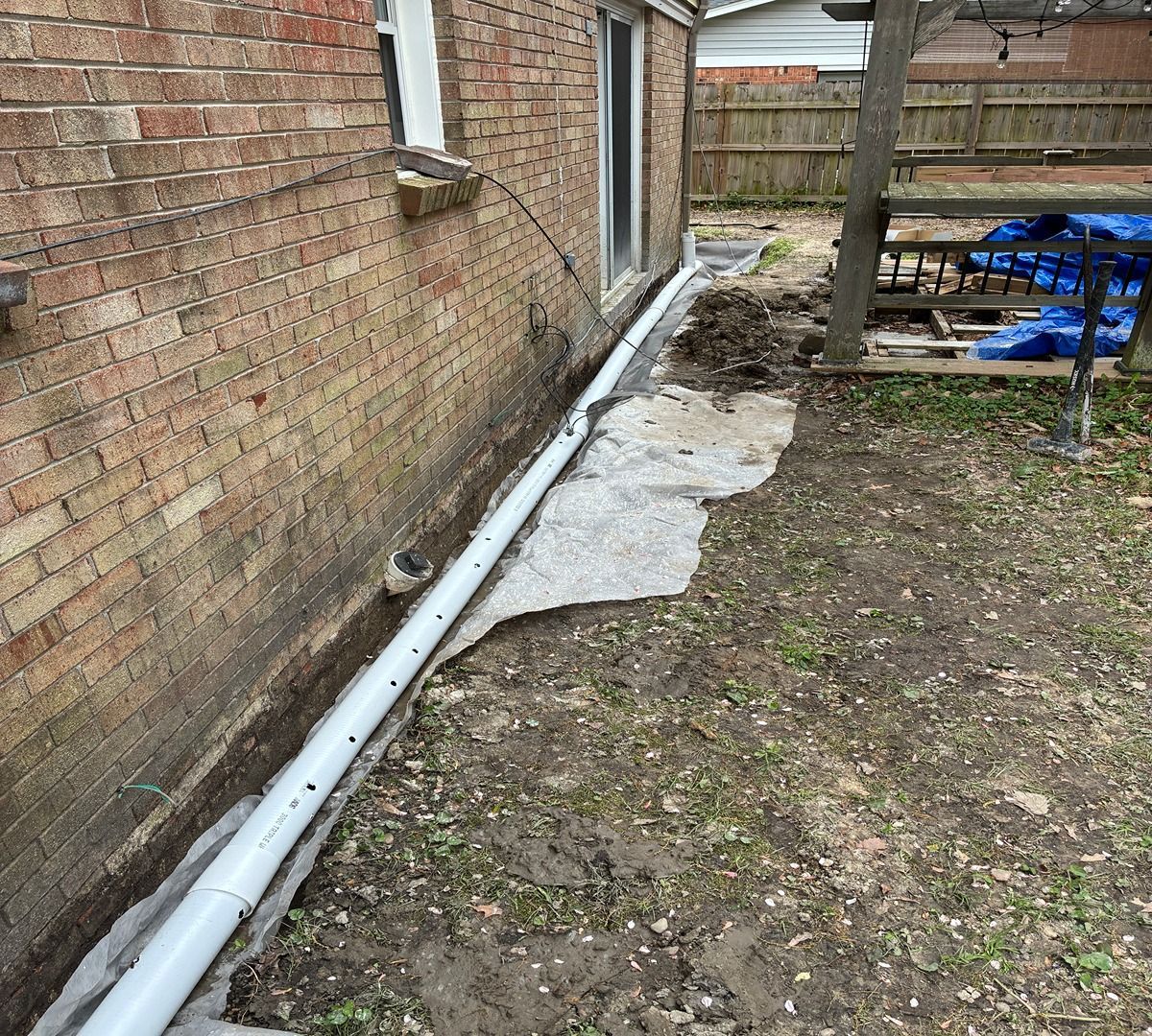 Side yard beside brick house with a white drainage pipe along the wall and patches of concrete on the ground
