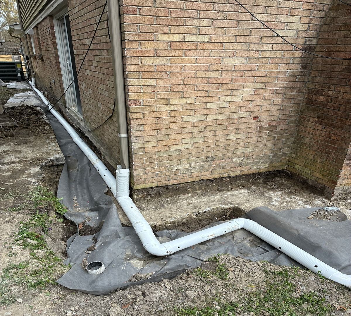 White drainpipe runs along the base of a brick wall over black landscape fabric in a narrow side yard.