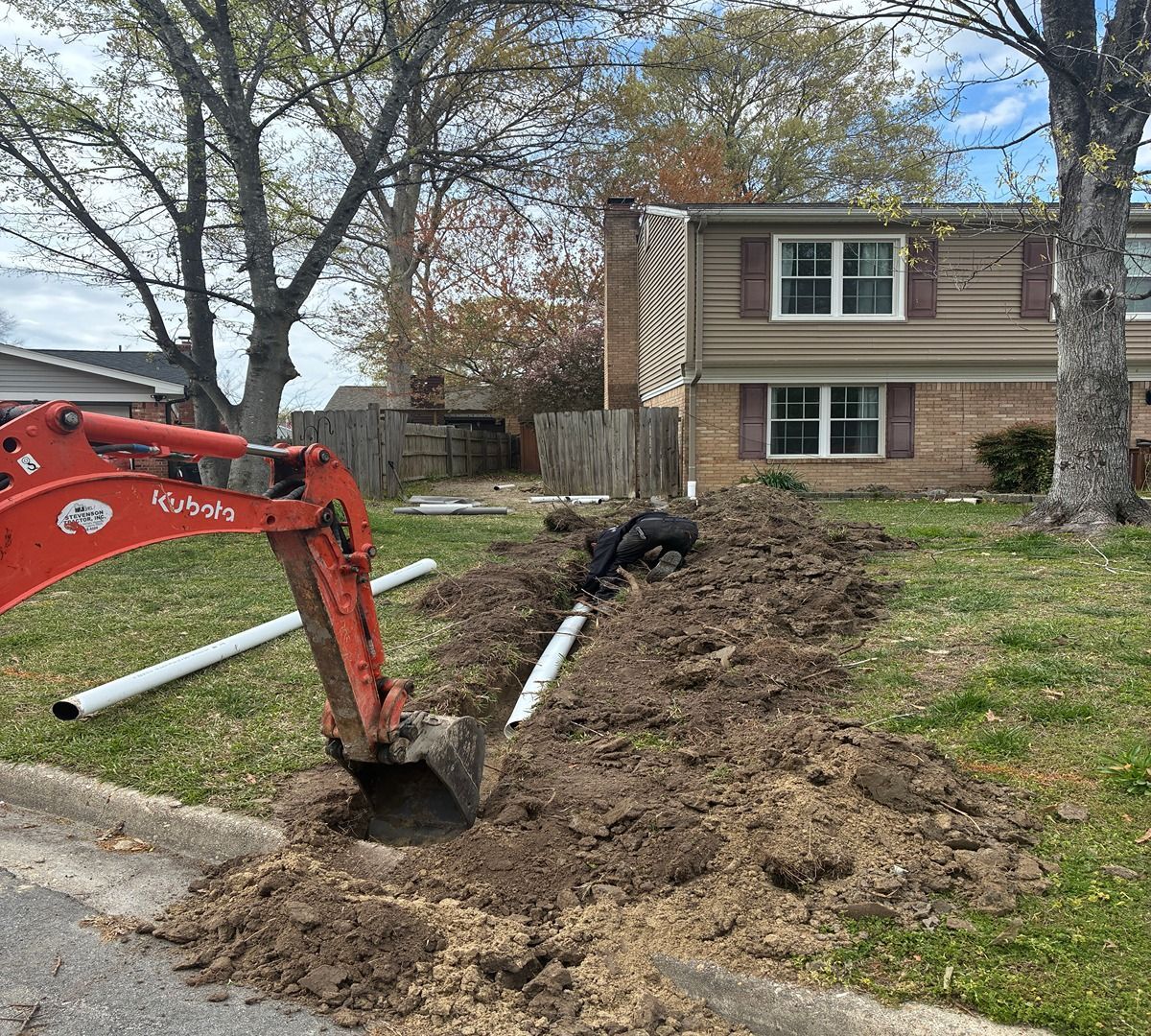 Orange excavator digging a trench beside a suburban house with a worker nearby