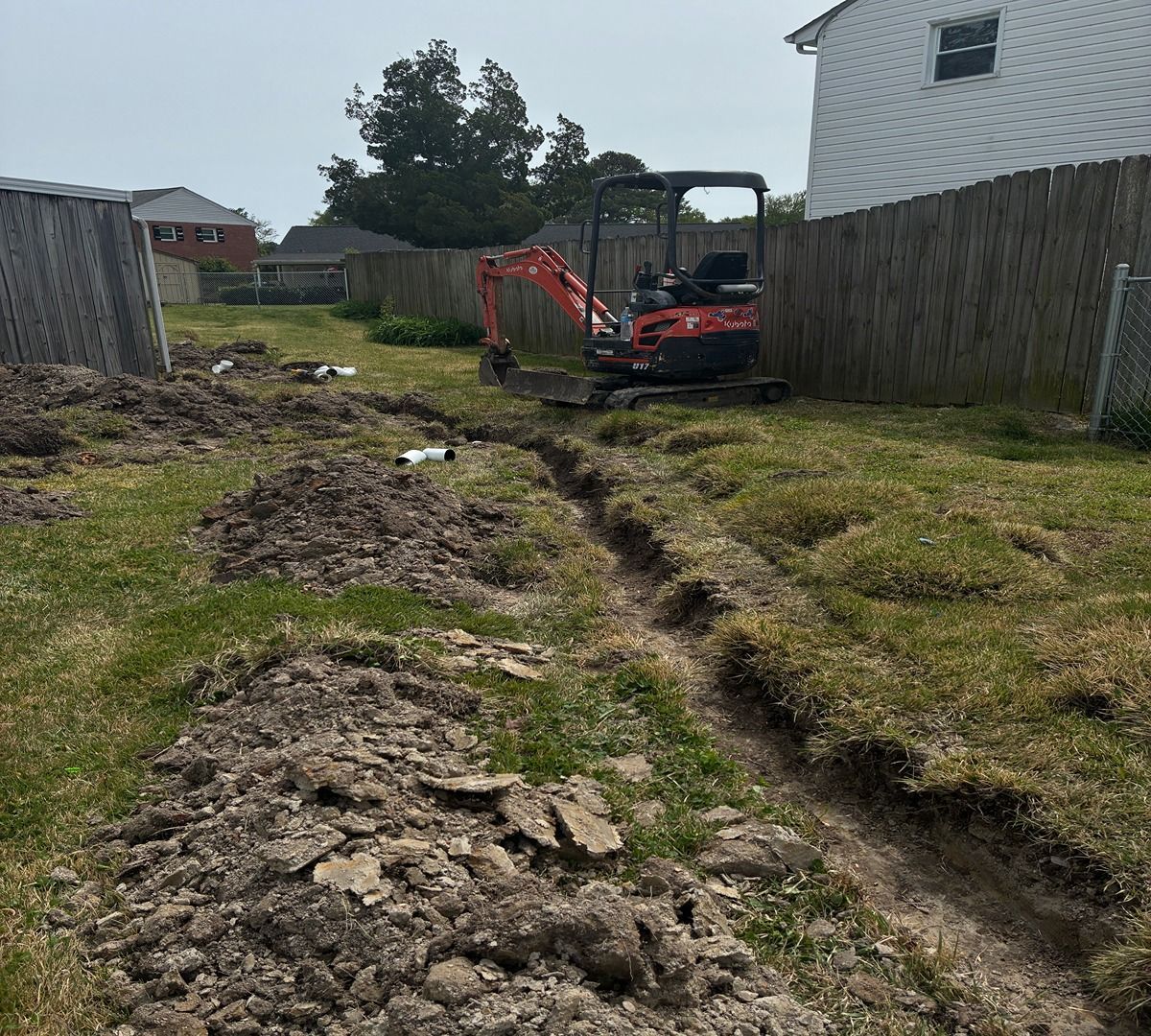 Mini excavator trenching a grassy backyard beside a house and fence.