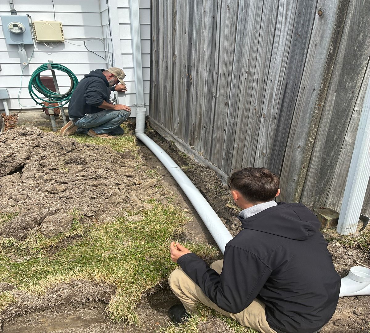 Two workers inspect a white drainage pipe along a wooden fence beside a house.