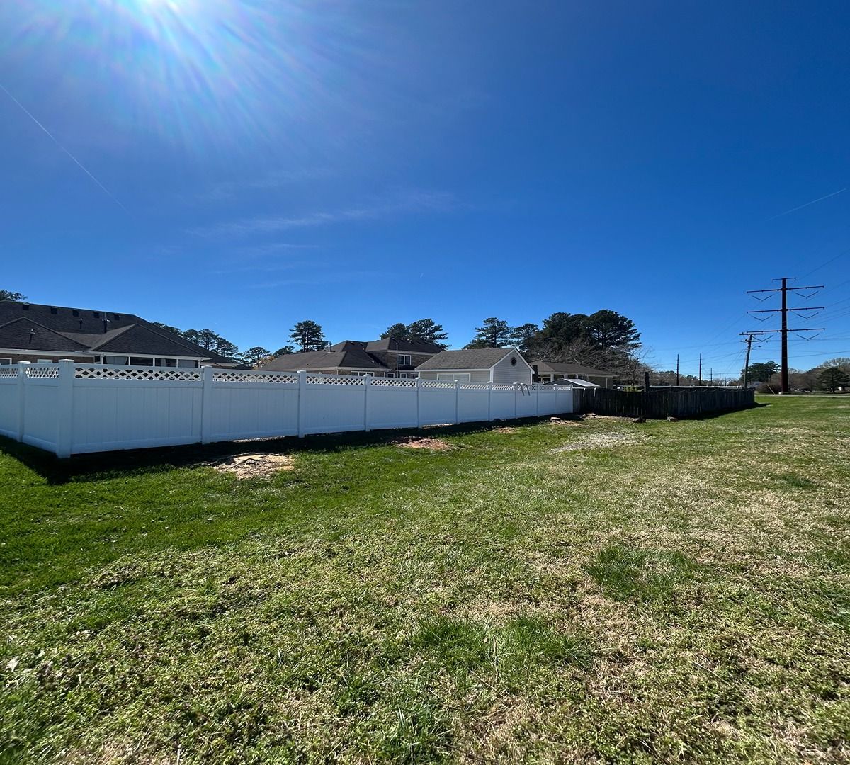 Grassy lot beside a white fence under a bright blue sky, with houses and power lines in the distance