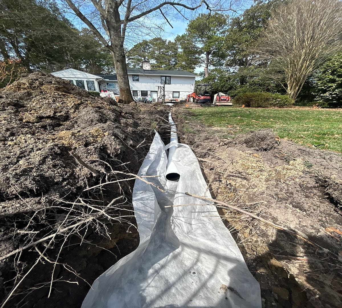 Drainage pipe lined with white fabric trench in a yard, with a house and trees in the background