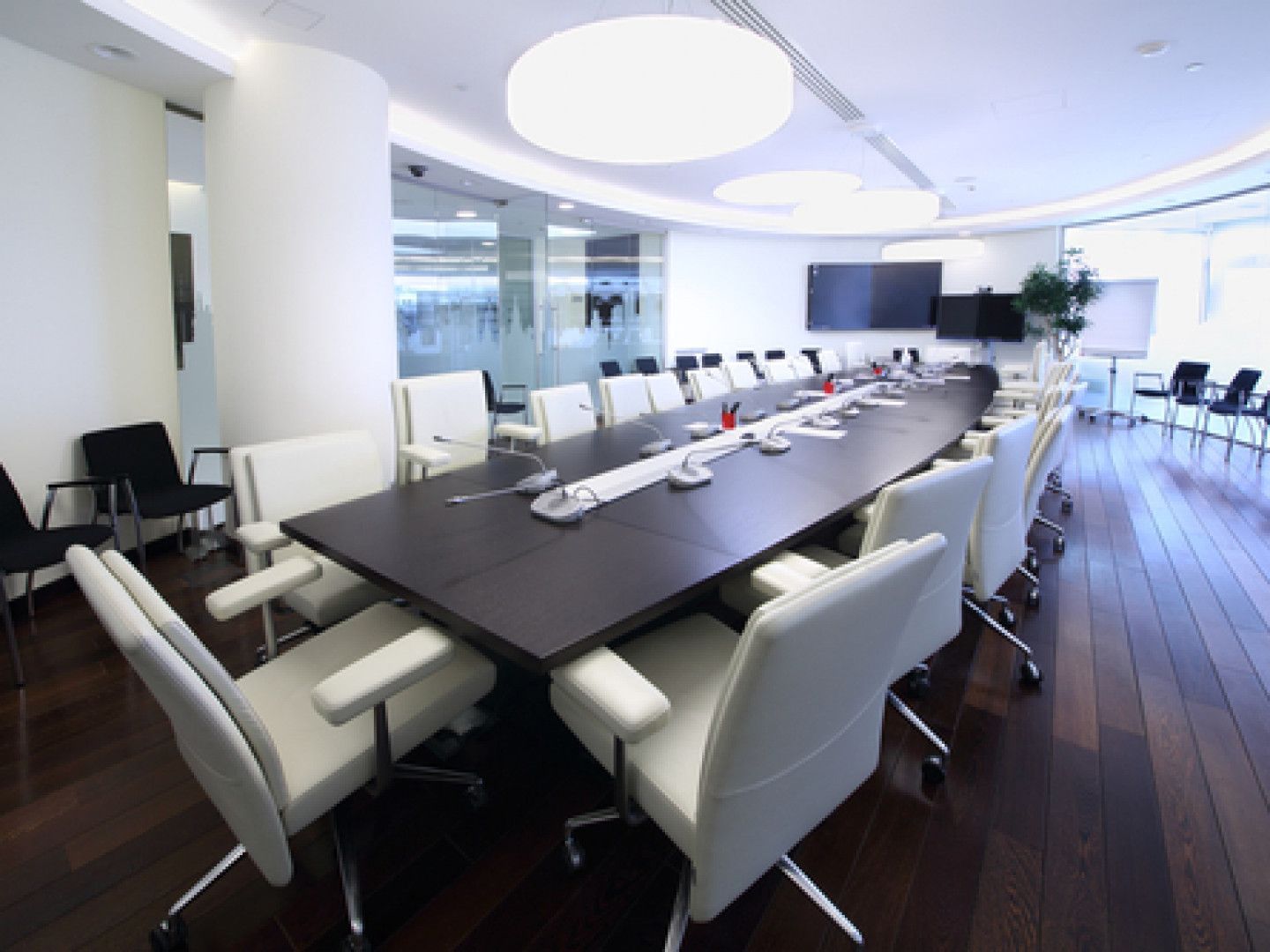 Conference room with long black table, white and black chairs, two screens, and a round overhead light.