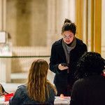 A person in a black jacket looks down at their phone while standing near two individuals at a table in a bright room.