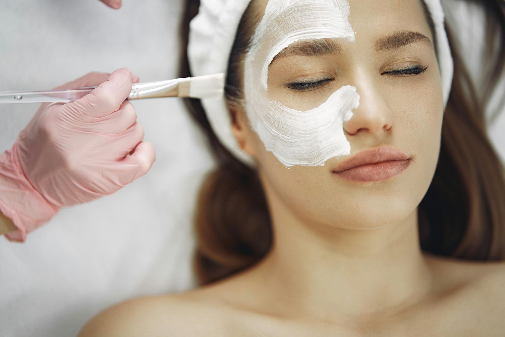 Person receiving facial mask treatment. White mask applied with a brush by a gloved hand.