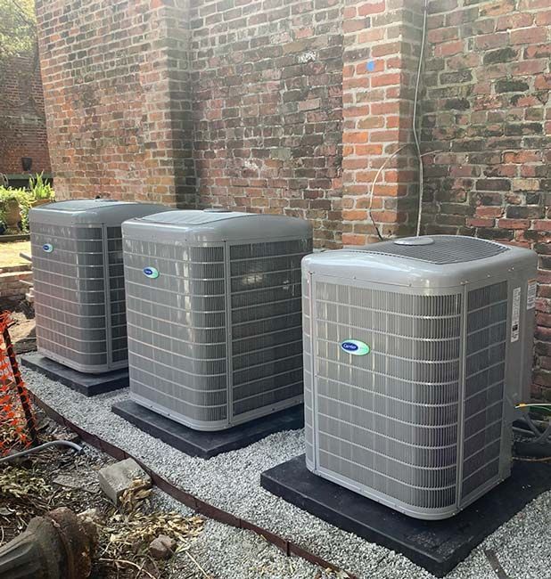 Three gray outdoor air conditioning units on pads beside a brick wall