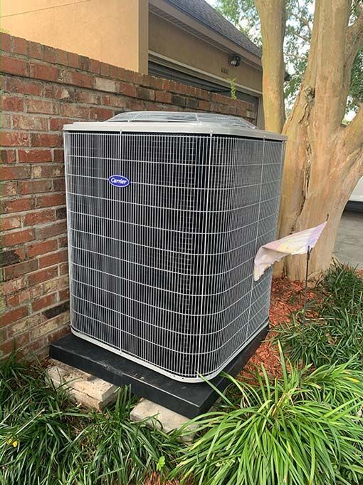 A gray Carrier outdoor air conditioning unit sits on a black pad next to a brick wall and a tree.