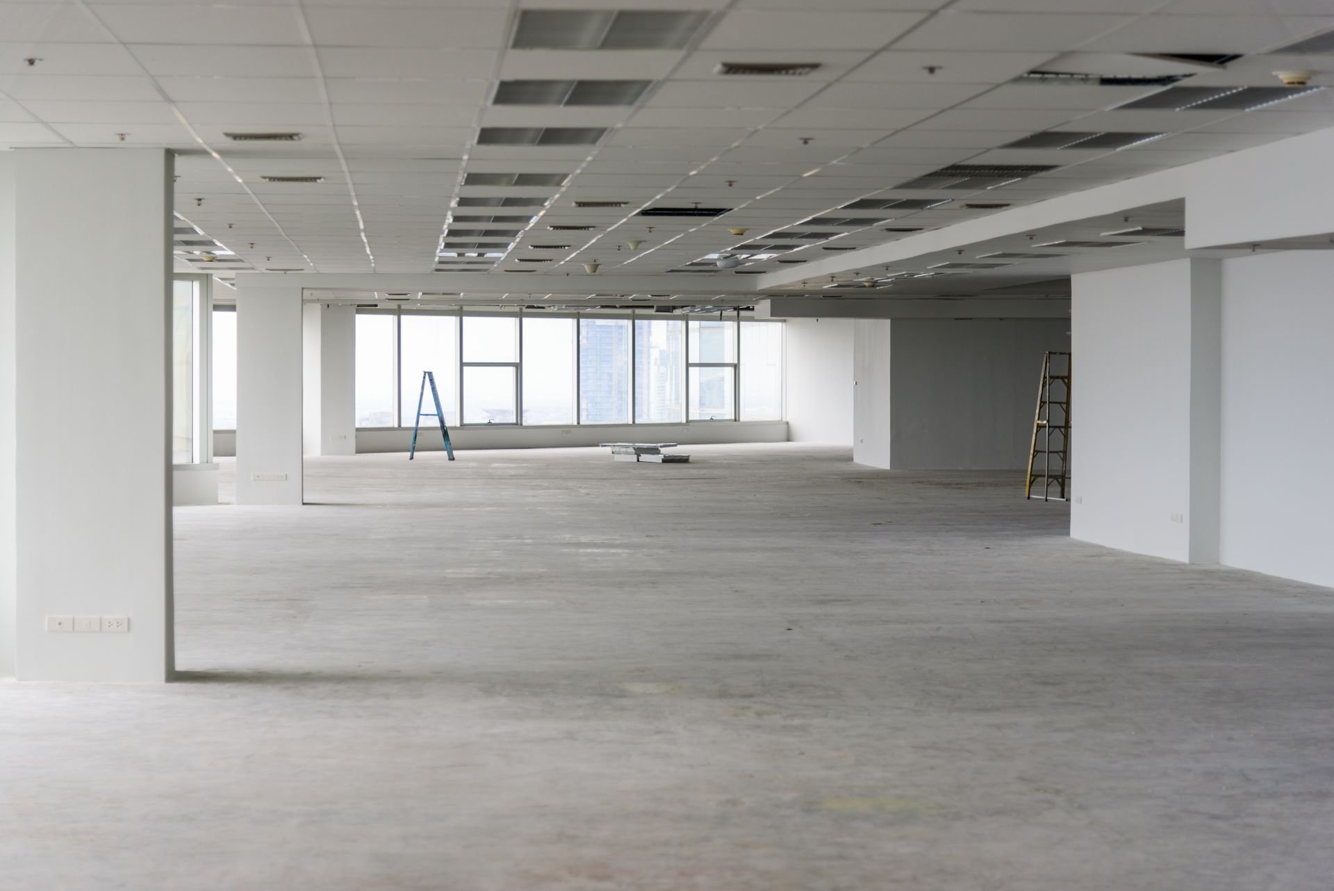 Espace de bureau vide avec moquette grise, murs blancs et grandes fenêtres sous un faux plafond