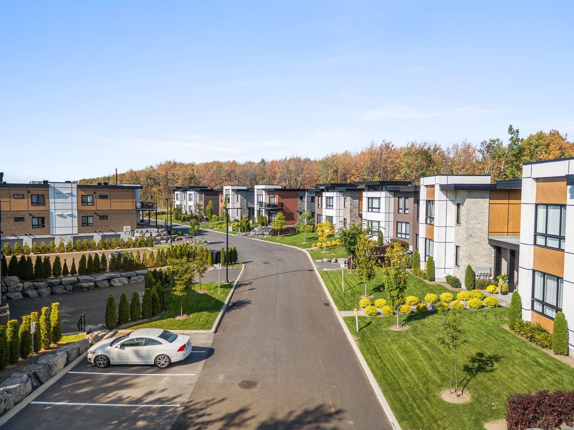 Modern apartment complex with a central road, parked cars, and autumn trees under a clear sky