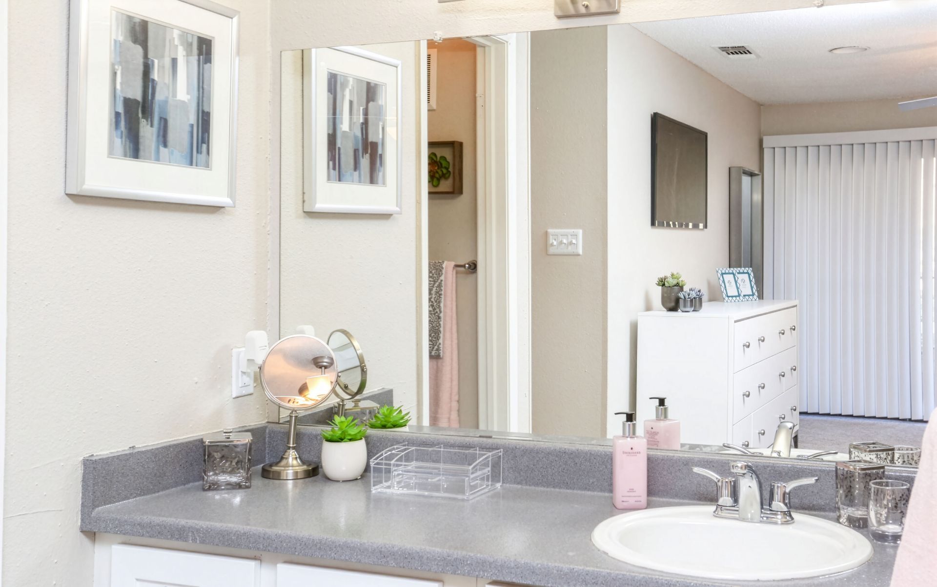 Bright bathroom vanity with gray countertop, sink, mirror, framed art, and a glimpse of the bedroom beyond
