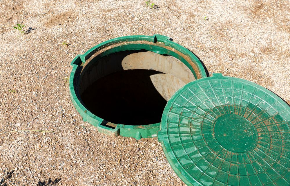 Green open manhole with cover on gravel ground.