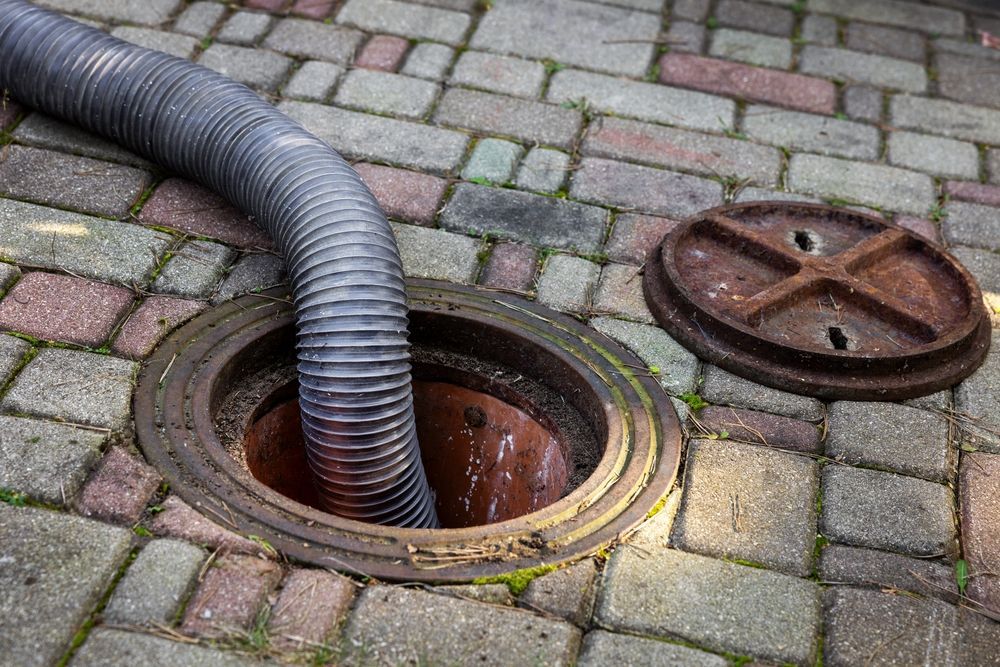 A corrugated hose extends into an open sewer manhole on a brick-paved surface; the cover is next to it.