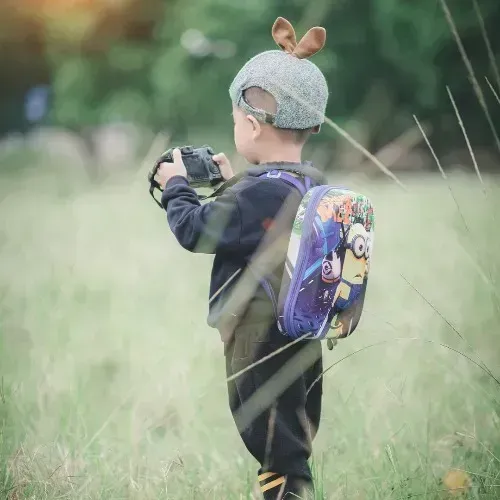 Child in a gray knit hat with bunny ears holding a camera in a grassy field, wearing a colorful backpack.