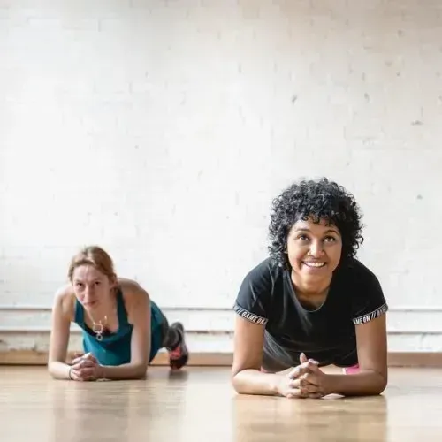 Two people doing a forearm plank on a wooden floor in a bright studio, smiling at the camera.