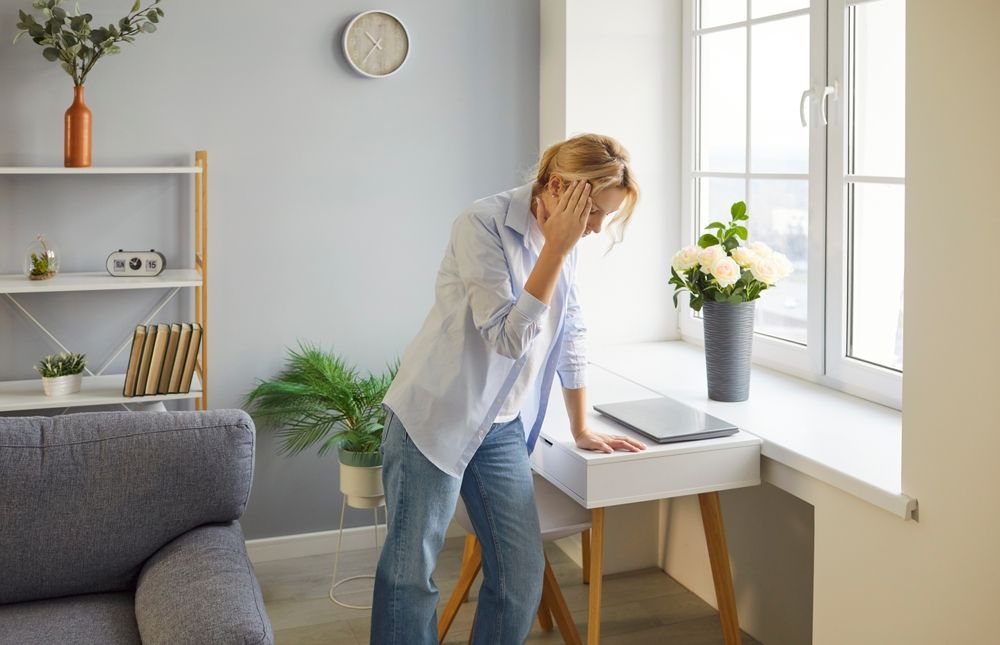 Person holding head in a bright home office beside a window, with a laptop on the desk.