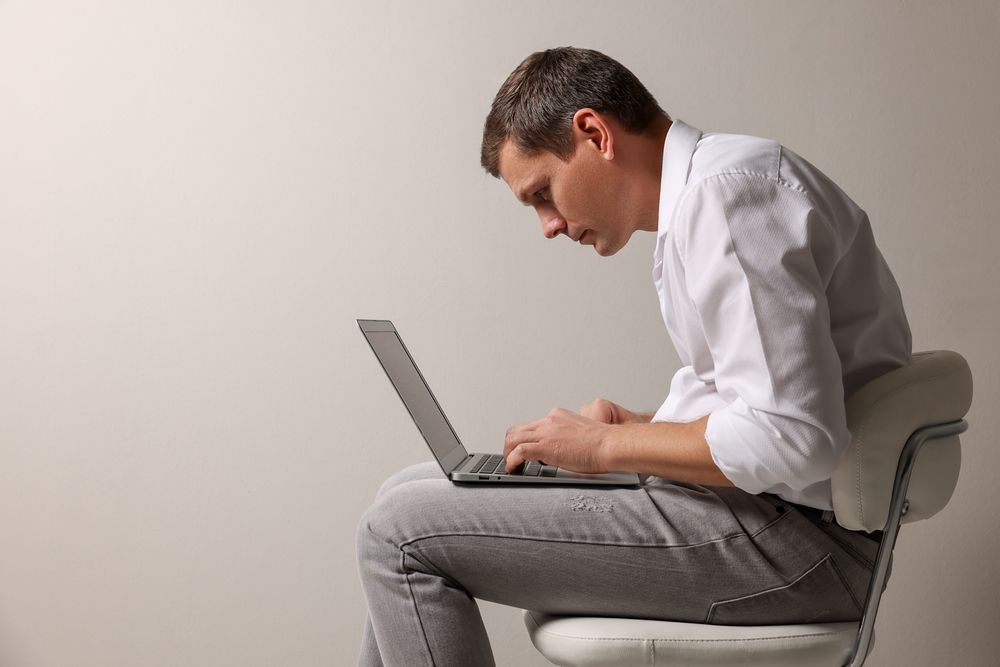 Man in a white shirt typing on a laptop while seated on a chair against a plain background