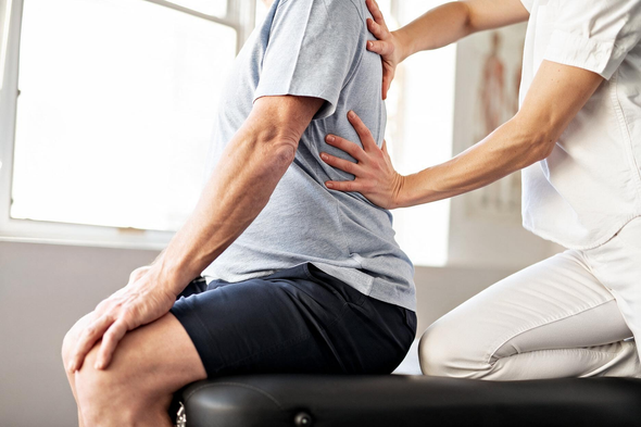 Physical therapist treating a seated patient’s back and leg on a therapy table by a window