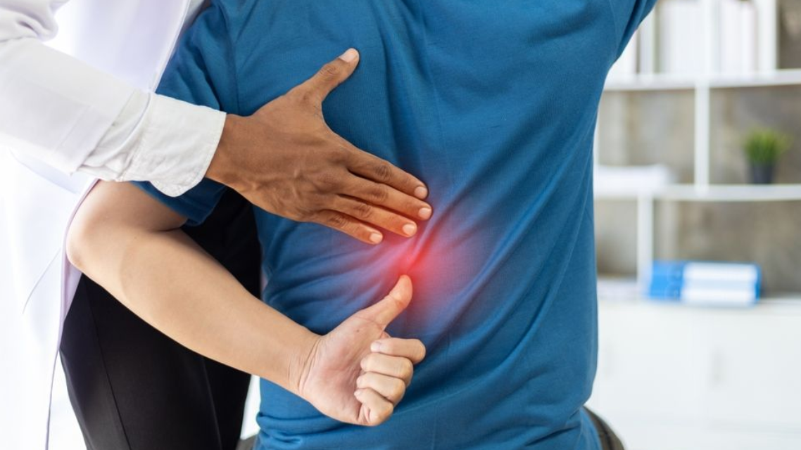 Person clutching lower back while a hand presses the side, suggesting back pain in a bright room