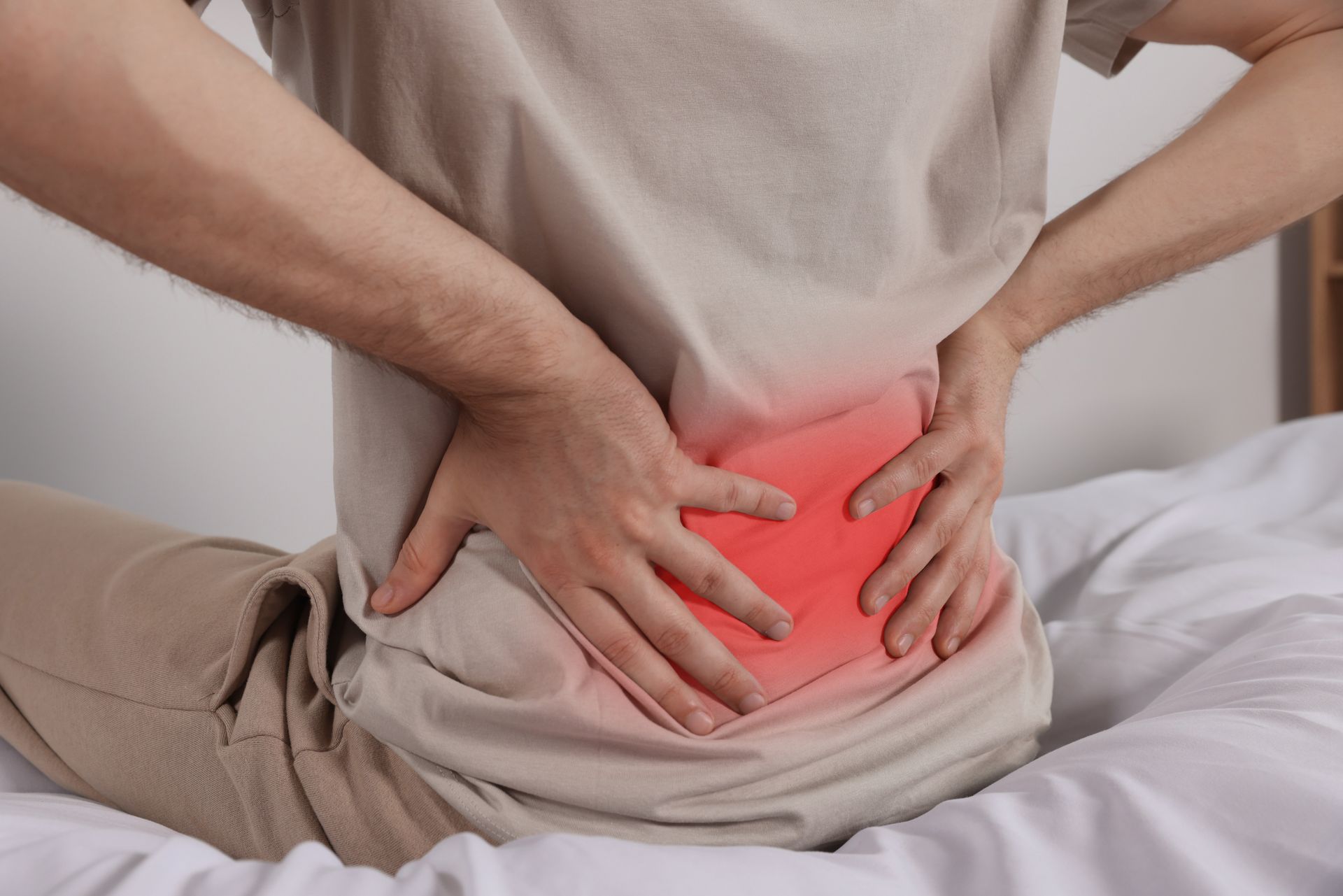 Person clutching lower back while sitting on a bed, with red pain highlight over the lower spine.