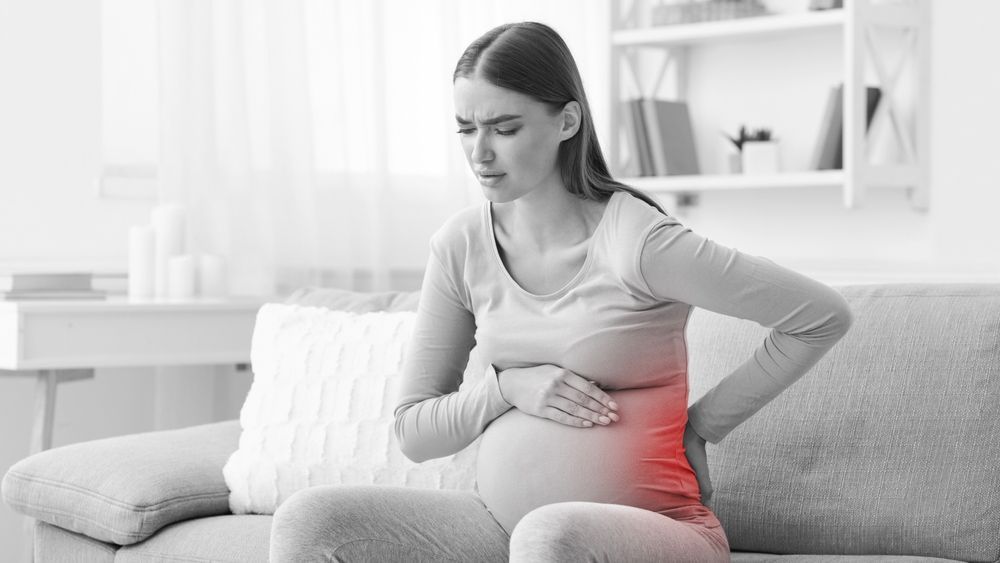 Woman on a couch holding her abdomen and lower back, appearing in pain at home.