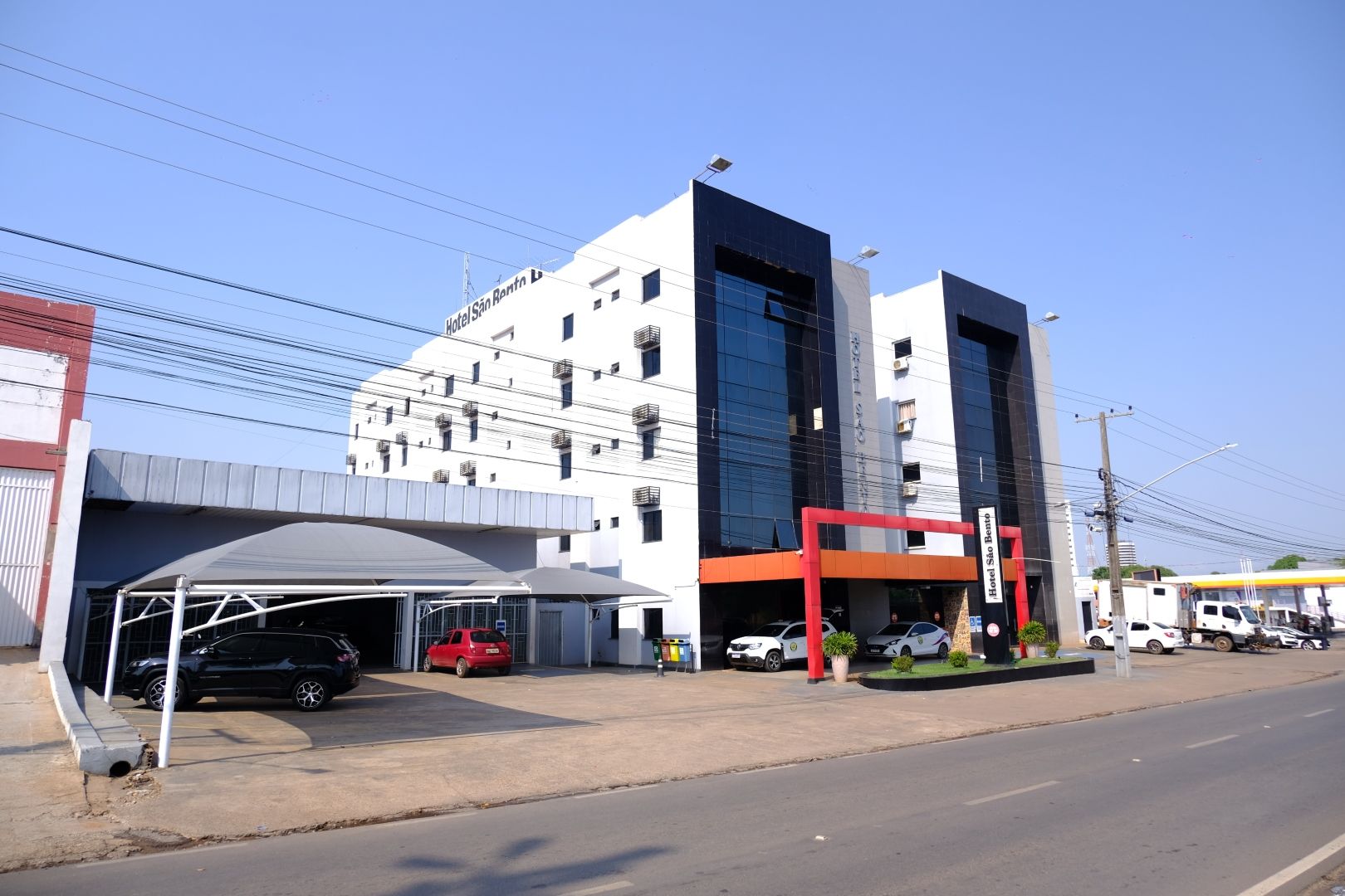 Prédio moderno branco com detalhes em preto, estacionamento na frente, carros estacionados, dia ensolarado.