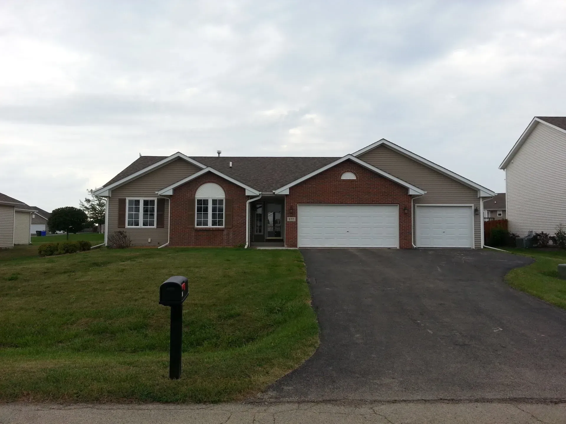 Single-story house with brick accents, two-car garage, and asphalt driveway under a cloudy sky.