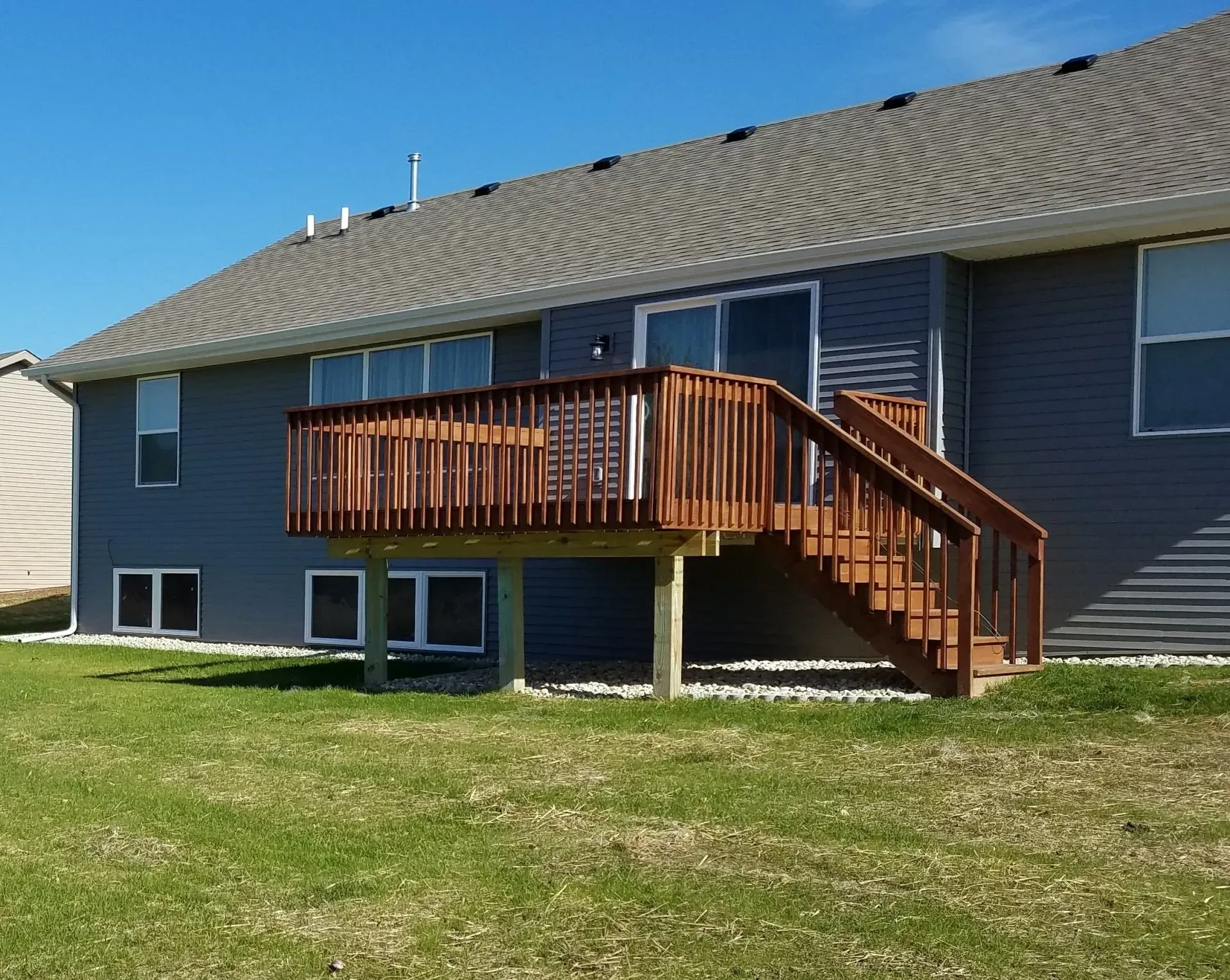 Wooden deck with stairs attached to a house with gray siding and a green lawn.