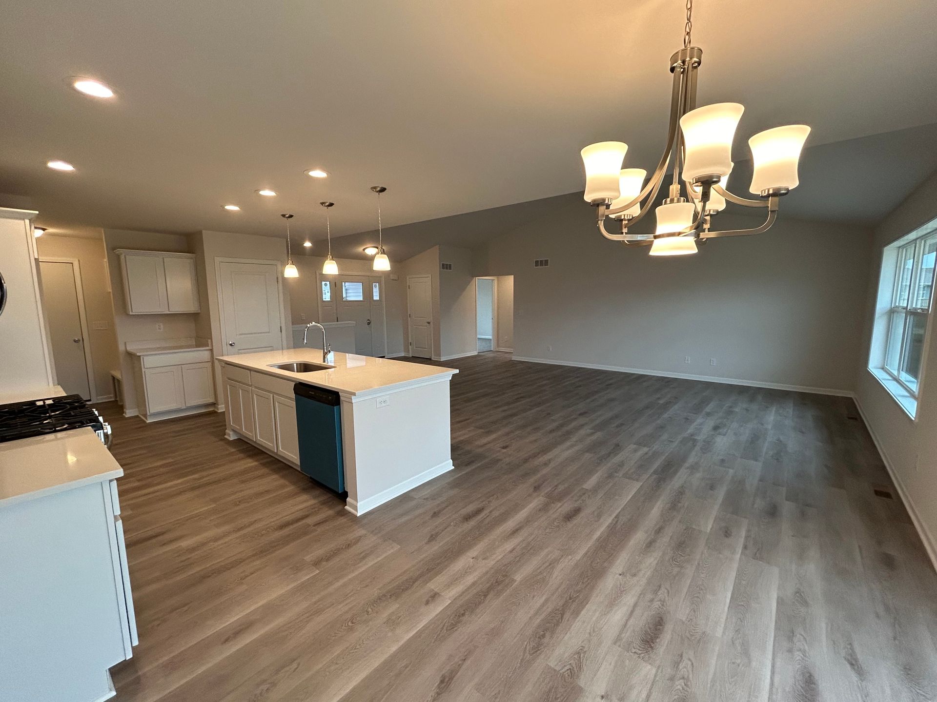 Open kitchen and living room with white cabinets, island, gray flooring, chandelier, and natural light.
