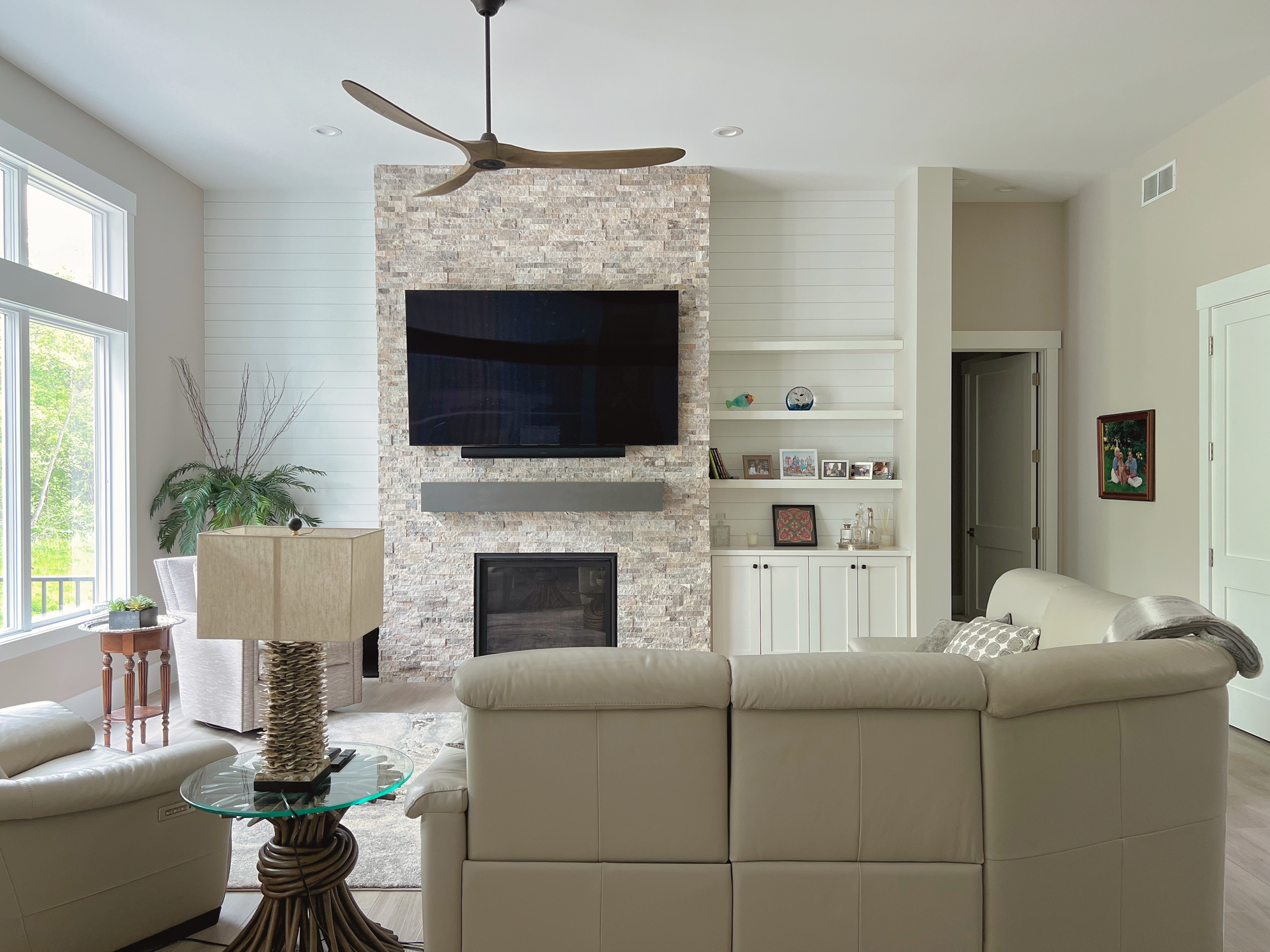 Living room with fireplace, TV, and light-colored sectional sofa. Beige and white tones with a wooden ceiling fan.