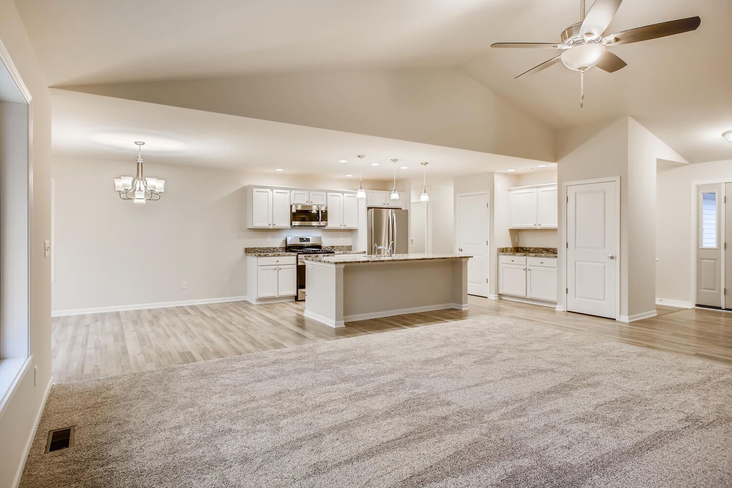 Open-concept living space with kitchen. Neutral tones: white walls, cabinets, island; gray carpet, wood floor.