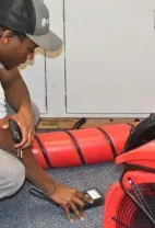 A person in a cap and gray shirt uses a handheld moisture meter on a carpet near a red industrial air mover and ducting.