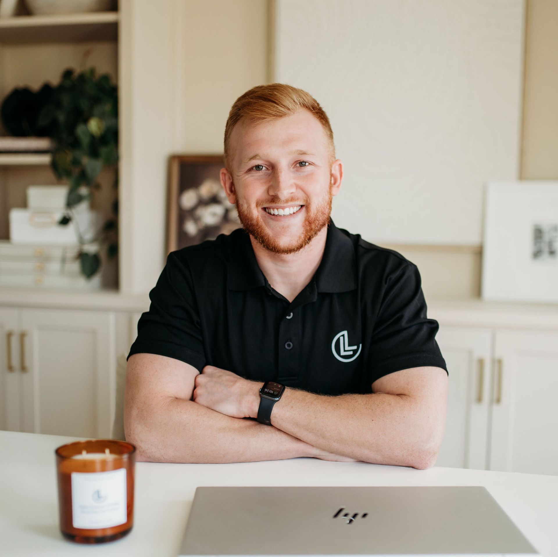 Smiling person in a black polo seated at a desk with a laptop and candle in a bright room