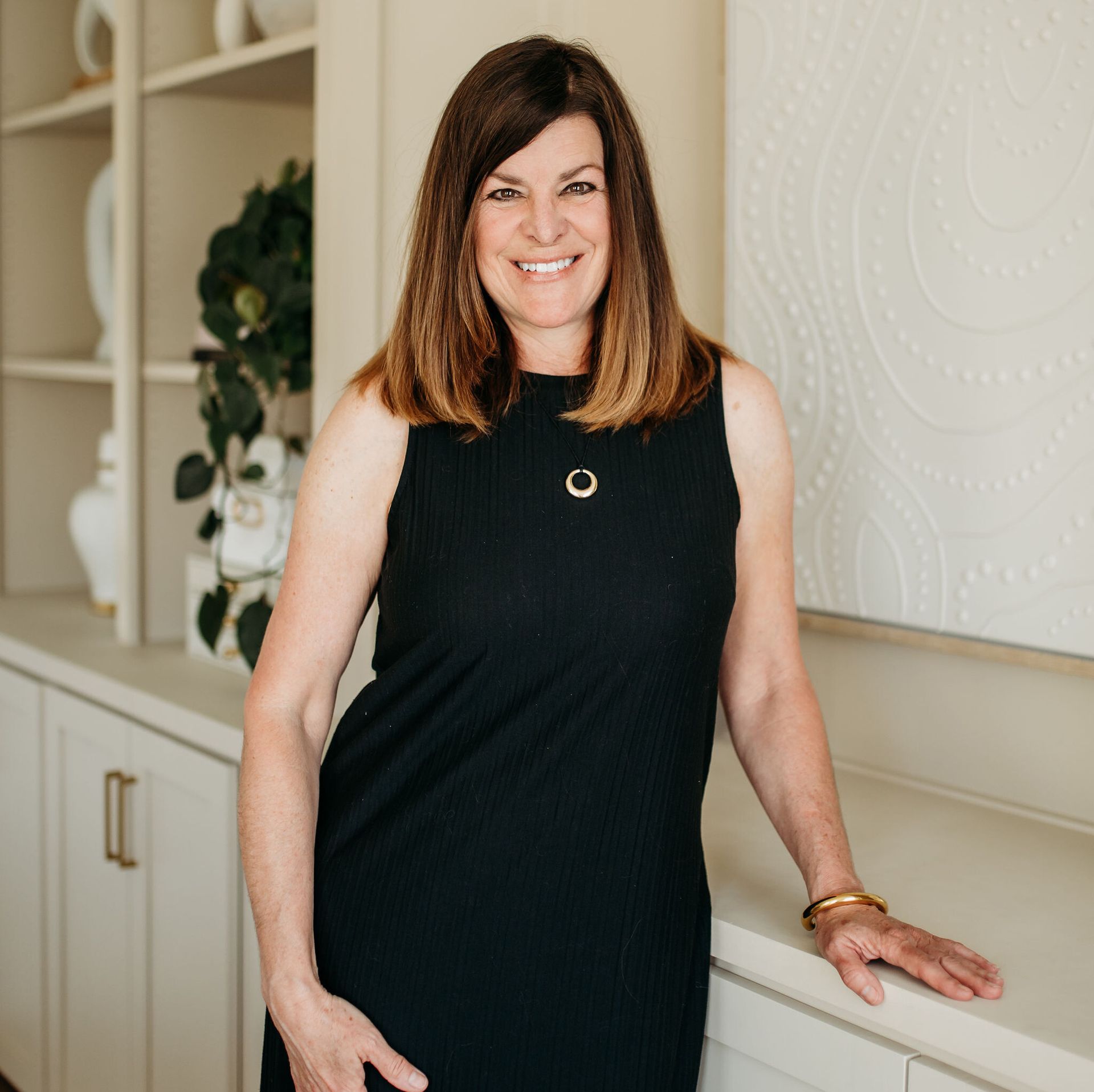 Woman in a black sleeveless dress leaning on a built-in shelf, smiling in a bright interior.