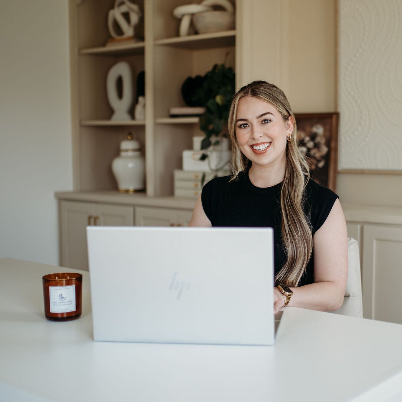 Woman smiling behind a laptop at a white desk in a bright home office