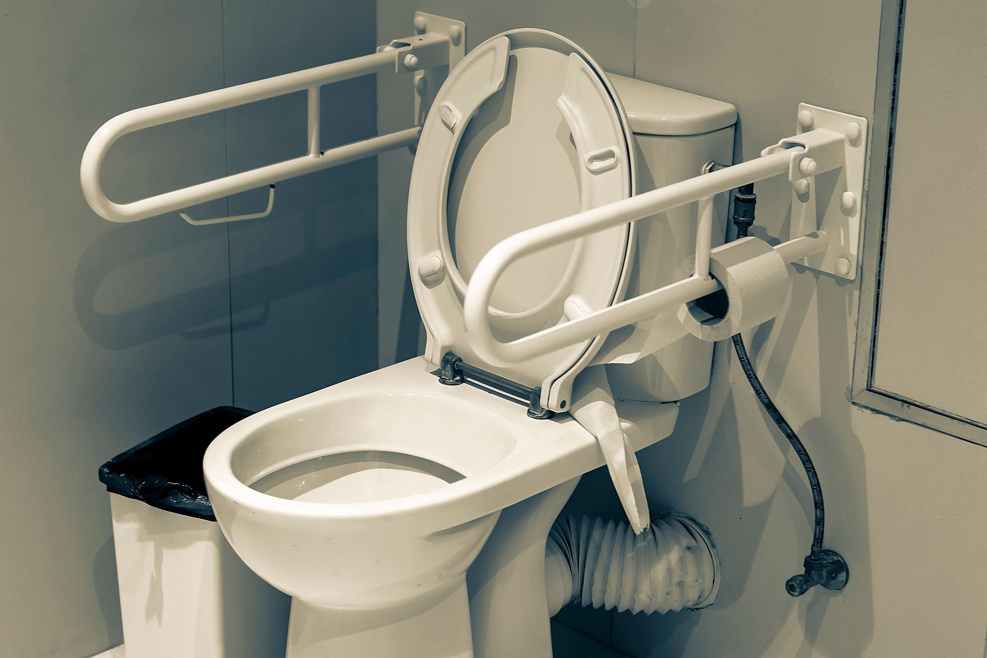 A toilet with a handicapped handle and a trash can next to it