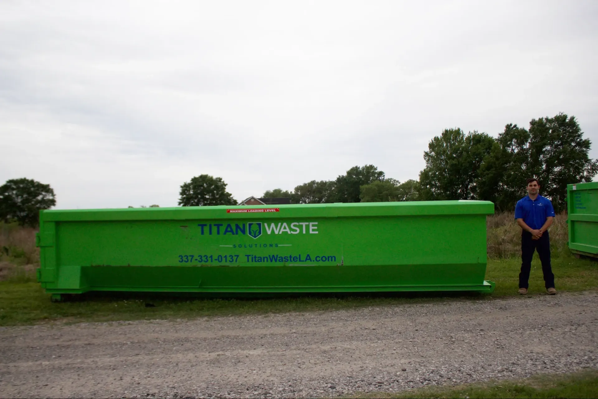 A man is standing in front of a large green dumpster.